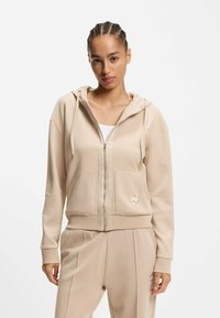 Sudadera con capucha beige con cremallera en tela suave, con un bolsillo frontal y detalle de logo. Cuenta con capucha ajustable con cordón y un ajuste relajado, combinada con pantalones a juego.