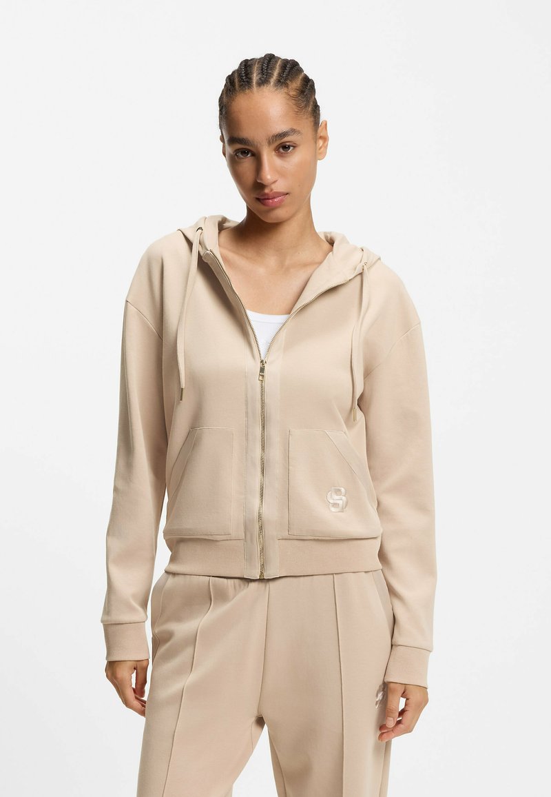 Sudadera con capucha beige con cremallera en tela suave, con un bolsillo frontal y detalle de logo. Cuenta con capucha ajustable con cordón y un ajuste relajado, combinada con pantalones a juego.