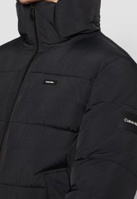 Czarna pikowana kurtka z wysokim kołnierzem, z przednim suwakiem i naszywkami z logo Calvin Klein na piersi i rękawie. Gładka, lekka tkanina.