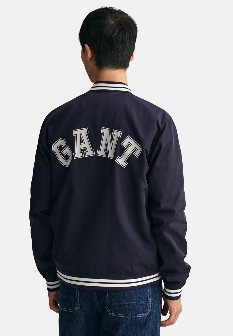Marine-Bomberjacke mit weiß gestreiftem Kragen und Bündchen. "GANT" in großen grauen Buchstaben auf dem Rücken aufgedruckt. Glattes Material, lässiges Design.