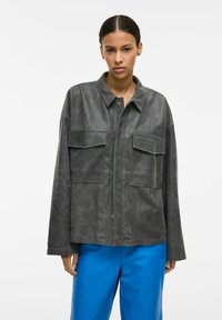 Szara zamszowa kurtka o luźnym kroju, z zapięciem na guziki, dużymi kieszeniami na piersi i lekko usztywnionymi ramionami, zestawiona z niebieskimi spodniami.