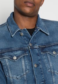 Chaqueta de mezclilla de color azul claro con un diseño clásico, que presenta dos bolsillos en el pecho, herrajes plateados y acentos de costura en color naranja.