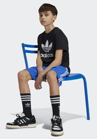 T-shirt noire Adidas avec logo blanc, shorts bleus, chaussettes à rayures noires et blanches, baskets noires avec accents blancs, assis sur une chaise bleue.