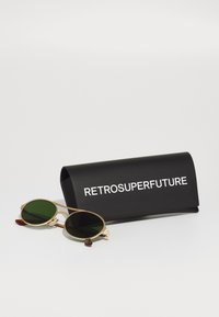 Gafas de sol redondas con lentes verdes, marco de metal dorado y acentos marrones en las patillas, junto a un estuche negro con el texto blanco "RETROSUPERFUTURE".