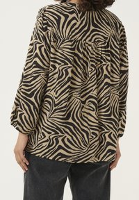 Blouse beige à rayures zèbre noires, dotée d'un dos froncé, de manches trois-quarts et d'une coupe décontractée. Associée à un pantalon foncé.