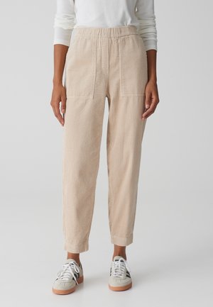 Woman wearing beige corduroy pants with elastic waistband and white sneakers, standing against a plain light background.