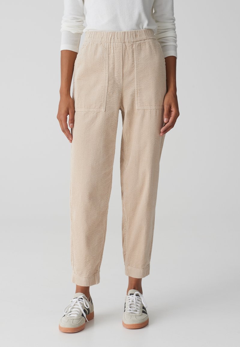 Femme portant un pantalon en velours côtelé beige avec une ceinture élastique et des baskets blanches, debout devant un fond clair uni.