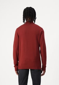 Jeune homme avec des dreadlocks mi-longs portant un pull à manches longues rouge foncé et un pantalon sombre, vu de dos sur un fond blanc.