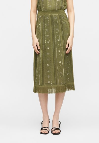 Μίντι φούστα λαδί με κάθετα σχέδια και τελείωμα με κρόσσια, φορεμένη με μαύρα σανδάλια με ψηλό τακούνι και λουράκια.