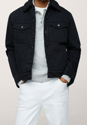 Homme portant une veste en jean noire boutonnée sur un t-shirt henley gris clair et un pantalon blanc, mains dans les poches, sur un fond uni.
