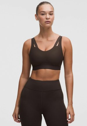 Donna che indossa un reggiseno sportivo marrone scuro e leggings a vita alta, in piedi davanti a uno sfondo grigio chiaro uniforme con capelli raccolti all’indietro.