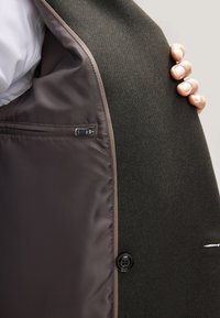 Donkere wollen jas met een gestructureerde buitenstof. Aan de binnenkant een gladde bruine voering en een ritssluitingzak. Zwarte knoop aan de voorkant.