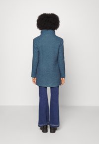 Manteau en laine teal avec un col haut, des manches longues et une coupe droite, associé à un jean en denim évasé et des chaussures noires massives.