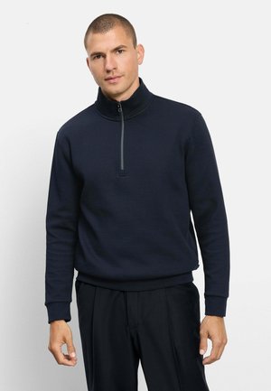 Uomo che indossa un maglione blu navy scuro con zip e collo alto, in piedi contro uno sfondo chiaro e semplice.