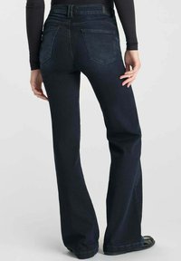 Persoon in donkerblauwe flare jeans en een zwart langemouwen shirt, staand met één hand op de heup.