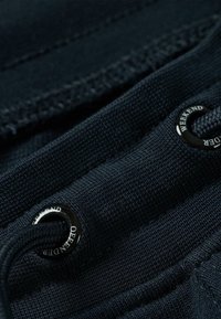 Námornícky modrá textúrovaná tkanina s viditeľným šitím. Obsahuje čierne kovové očká a šnúrku na nastavenie. Vzory zahŕňajú diagonálne rebrovanie.