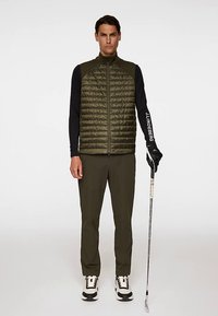 Gilet imbottito verde oliva indossato sopra una maglia nera a maniche lunghe, abbinato a pantaloni color oliva. L'individuo tiene in mano una mazza da golf, con scarpe da ginnastica nere e bianche visibili.