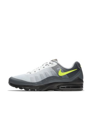 Zapatilla deportiva Nike con parte superior de malla blanca, detalles en gris, suela negra y un logo amarillo vibrante. El diseño incluye un talón acolchado para mayor soporte.