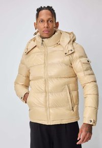 Homme aux cheveux tressés portant une veste matelassée beige clair avec poches zippées et poignets à boutons-pression, debout devant un fond uni.