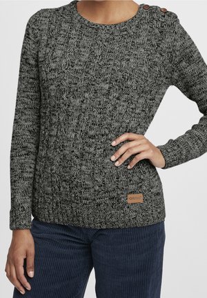 Femme portant un pull gris en maille torsadée avec des boutons aux épaules et un petit patch logo, associé à un pantalon en velours côtelé bleu foncé.