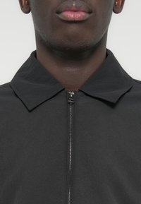 Μαύρο μπουφάν με φερμουάρ, διαθέτει επίπεδη γιακά και λεία υφή, με καθαρό, μινιμαλιστικό σχέδιο και λειτουργική λεπτομέρεια φερμουάρ.