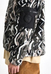 Fleece jacket with an abstract black, white, and tan pattern; features a zippered pocket and a textured black patch on the sleeve.