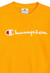 T-shirt in cotone giallo con colletto rotondo, caratterizzato da un logo "Champion" ricamato in bianco e un piccolo dettaglio rosso all'interno del logo.