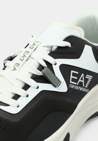 Közeli kép fekete-fehér tornacipőről fehér fűzőkkel, zöld fűzővégződésekkel és az oldalán "EA7 Emporio Armani" logóval.