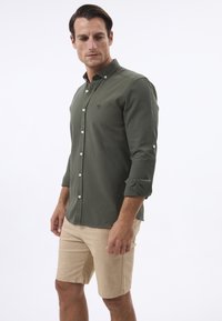 Chemise boutonnée vert olive à manches longues, avec un petit logo, assortie à un short beige clair en tissu texturé.