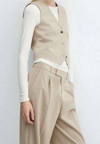 Beżowa kamizelka o dopasowanym kroju z konstrukcyjnym designem, z trzema ciemnymi guzikami i dekoltem w kształcie litery V. Połączona z beżowymi spodniami z wysokim stanem i plisy.
