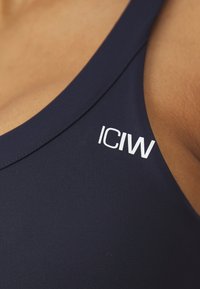 Tamsiai mėlynas sportinis liemenėlis, pagamintas iš lygaus audinio, su lenktos nugaros dizainu ir baltu "ICIW" logotipu viršutiniame kairiajame kampe.