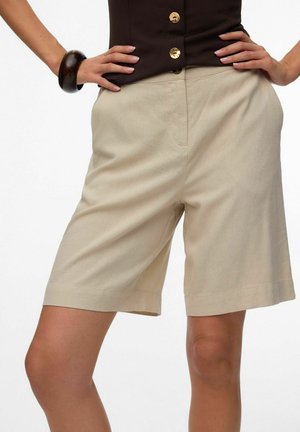 Mujer que lleva pantalones cortos beige hasta la rodilla y una blusa oscura con botones dorados, manos apoyadas en las caderas, y una pulsera oscura en la muñeca izquierda.