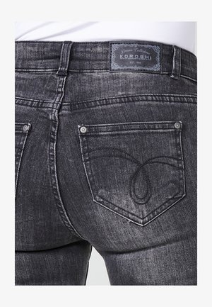 Jeans in denim grigio con un'etichetta che presenta la scritta "KOROSHI" e una progettazione distintiva delle tasche con cuciture curve. Texture dettagliata e vestibilità visibile.