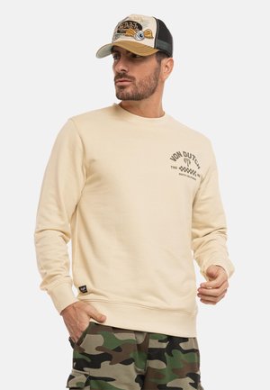 Homme portant un sweat-shirt beige Von Dutch, un pantalon cargo camouflage et une casquette de baseball beige et noire Von Dutch, regardant sur le côté.