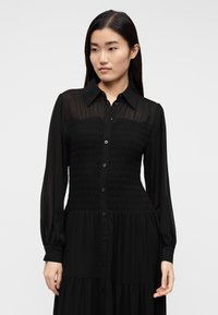 Čierne košeľové šaty s nariaseným živôtikom, dlhými priehľadnými rukávmi a viacvrstvovou sukňou. Hladká textúra a klasický golier.