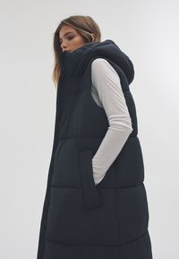 Tengerészkék, kapucnis, mellény, amely steppelt textúrával és két oldalzsebbel rendelkezik. Könnyű, hosszú ujjú felső fölött viselve.