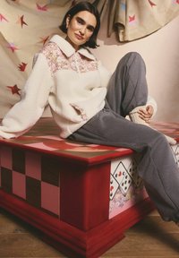 Pulover alb din fleece cu yoke cu model floral și guler mare, asortat cu pantaloni gri, așezat pe o masă cu model colorat.