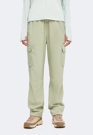 Persona que lleva pantalones cargo verde claro con bolsillos laterales, zapatos de senderismo beige y una camiseta ligera de manga larga, de pie frente a un fondo liso.