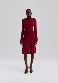 Κόκκινο ριμπ πλεκτό φόρεμα με ψηλό λαιμό, φουσκωτά μανίκια και βολάν στο τελείωμα. Συνδυάζεται με μαύρες μυτερές γόβες. Λείο υλικό.