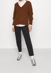 Pull-over oversize col en V couleur rouille en tissu doux, superposé sur une chemise blanche à col, associé à un pantalon noir coupe slim et des baskets argentées.