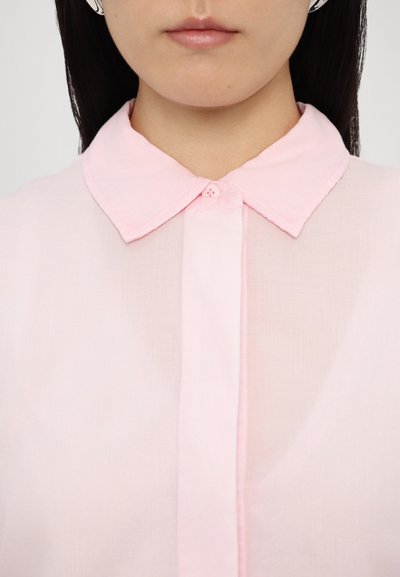 Chemise rose clair, semi-transparente, avec un col classique, dotée d'une fermeture à boutons et d'une texture subtile, mettant en avant la simplicité du design.