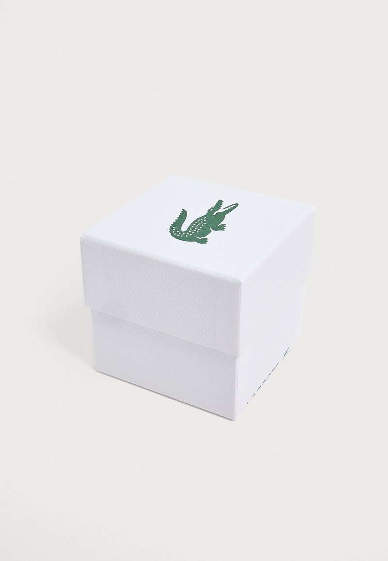 Boîte carrée blanche texturée avec un logo de crocodile vert centré sur le dessus, sur un fond clair uni.
