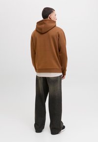 Mand iført en brun hættebluse, løse mørke jeans og sorte sneakers, står med ryggen til mod en simpel lys baggrund.