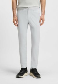 Pantaloni grigi chiari in un materiale texturizzato, vestibilità su misura, con zip e chiusura a bottone. Indossati con sneakers nere con suola bianca.