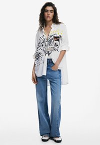 Weißes oversized Hemd mit schwarzen grafischen Designs und Text, kombiniert mit hellblauen Jeans und schwarz-weißen Schuhen.
