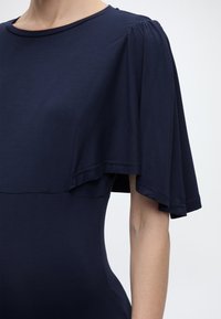 Μπλε ναυτικό φόρεμα με φαρδιές, αέρινες κοντές μανσέτες και στρογγυλή λαιμόκοψη, φορεμένο από ένα άτομο μπροστά σε απλό φόντο.