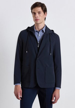 Man wearing a navy blazer with patch pockets over a zip-up hoodie and blue checkered shirt, standing against a plain light background.