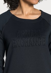 Osoba na sobě má tmavou mikinu s vyvýšeným textem "ARMANI EXCHANGE" na hrudi, zkrácenou pod nosem, takže jsou vidět krk a klíční kost.