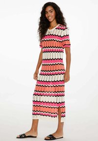 Pletené maxi šaty se zigzagovým vzorem v růžové, bílé a černé barvě; mají krátké rukávy a boční rozparek, kombinované s černými šlapkami.