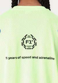 Grønn mesh trøye med rund hals. Bak er det en svart logo med "F1" og teksten "75 år med hastighet og adrenalin."